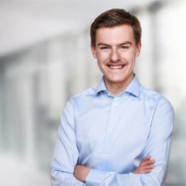 Junger Mann mit einem freundlichen Lächeln, der in einem hellblauen Hemd vor einem modernen, unscharfen Hintergrund steht.