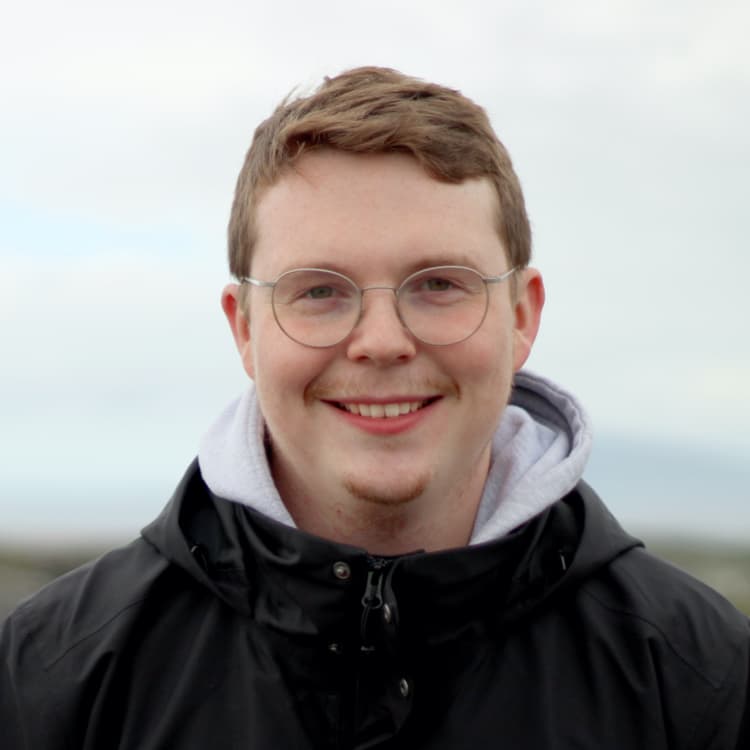 Junger Mann mit Brille und kurzem, lockigem Haar, lächelt in einer Outdoor-Jacke vor einem bewölkten Himmel.